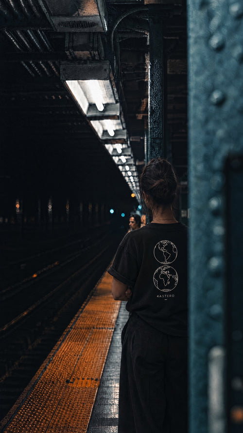 Woman with crossed arms on a dark subway platform in a black 4ASTERO tee with the Borderless globes back print
